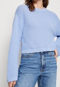 Žena oblečená v svetle modrém pleteném svetru s dlouhými rukávy a vysokým pasem modrých džín, stojící s jednou rukou na boku.