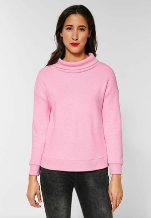 Maglione rosa a maglia con un collo alto e arrotolato e polsini a costine. Presenta una vestibilità rilassata e una texture liscia, abbinato a pantaloni scuri.
