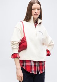 Νεαρή γυναίκα που φοράει λευκό φούτερ με φερμουάρ πάνω από κόκκινο καρό πουκάμισο, κρατώντας μια μικρή κόκκινη τσάντα στον ώμο, στέκεται με το ένα χέρι στη μέση.