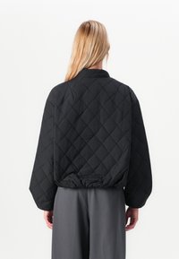 Femme aux longs cheveux blonds portant une veste matelassée noire et un pantalon gris, vue de dos sur un fond blanc uni.
