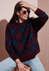 Pull en tricot avec un motif de losanges bordeaux et bleu marine, col rond côtelé et manches tombantes ; porté avec un jean bleu clair.