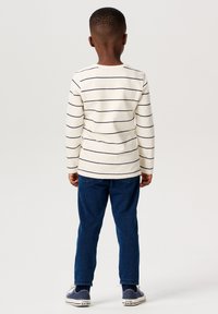 Langarm, crèmekleurig gestreept T-shirt met marineblauwe lijnen, gecombineerd met blauwe denim broek en marineblauwe schoenen, gezien vanuit de achterkant.