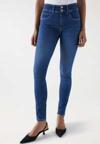 Vaqueros skinny de tiro alto en azul con cierre de dos botones, cinco bolsillos y una textura de denim suave. Combinados con tacones de tira negros.