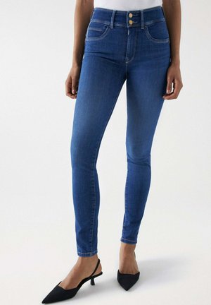 Vaqueros skinny de tiro alto en azul con cierre de dos botones, cinco bolsillos y una textura de denim suave. Combinados con tacones de tira negros.