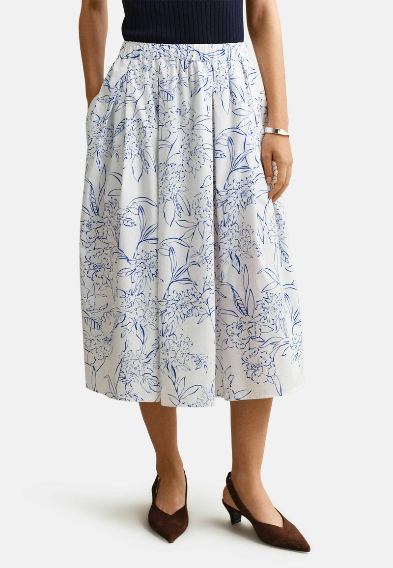 Frau trägt einen weißen Midirock mit blauem Blumenmuster, kombiniert mit braunen spitz zulaufenden Slingback-Absätzen und einem silbernen Armband.