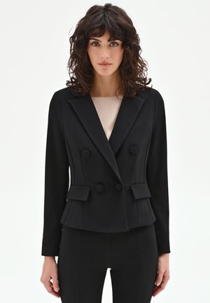 Donna con capelli ricci e scuri che indossa un blazer nero doppiopetto aderente e pantaloni abbinati, in piedi davanti a uno sfondo bianco semplice.