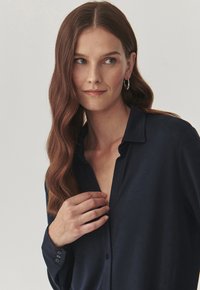 Donkerblauwe geknoopte blouse met een gladde textuur, een kraag en een licht opengeknoopte halslijn. Heeft geknoopte manchetten en een ontspannen pasvorm.
