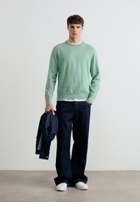 Jeune homme debout contre un mur blanc, portant un pull vert, un jean large foncé, des baskets blanches, tenant une veste noire dans sa main gauche.