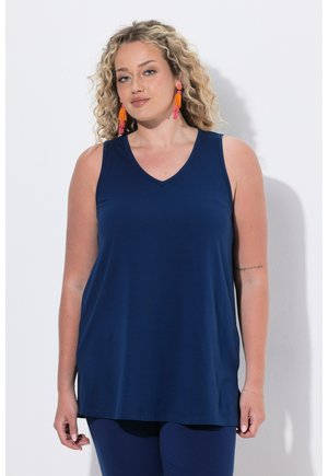 Frau mit lockigem blondem Haar, die ein marineblaues ärmelloses Oberteil und leuchtend orange-pinke Quastenohrringe vor einem schlichten hellen Hintergrund trägt.