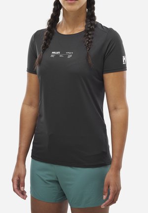 Femme aux cheveux tressés portant un t-shirt Millet noir à manches courtes et un short de sport bleu sarcelle sur un fond blanc uni.