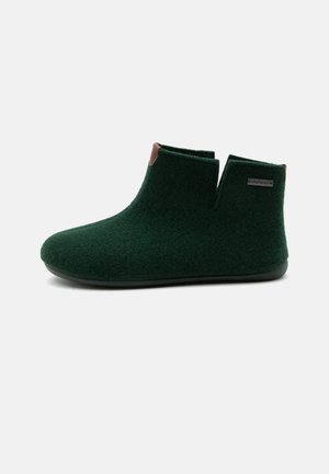 Pantoufle cheville en tissu doux vert foncé avec semelle noire et petit logo rectangulaire sur le côté sur un fond clair uni.