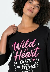 Černé bavlněné tričko s kulatým výstřihem a tříčtvrtečními rukávy. Na přední straně je růžový a bílý nápis: „Wild at Heart Crazy in Mind.“