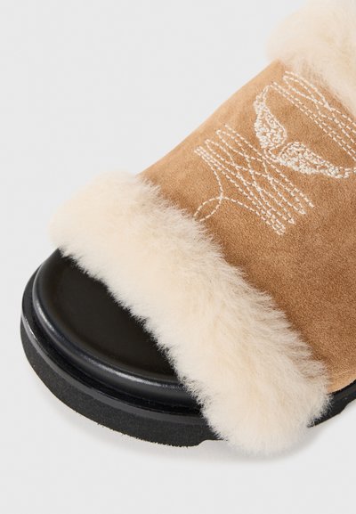 Braune Wildleder-Sandale mit flauschigem cremefarbenem Lammfellfutter, ausgestattet mit einer schwarzen Gummisohle und einem einfachen gestickten Design auf dem Schaft.