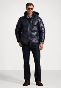 Mornarsko plava proćelava jakna s sjajnim završetkom, zatvaračem i kapuljačom. Nošena preko mornarskog džempera i uparena s tamnim trapericama i smeđim mokasinama.