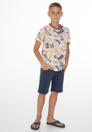 Junge steht mit den Händen in den Taschen, trägt ein buntes grafisches T-Shirt, dunkle knielange Shorts und Flip-Flops vor weißem Hintergrund.