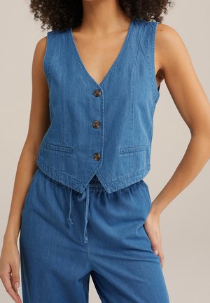 Vrouw draagt een mouwloos blauw button-down vest met V-hals en bijpassende broek met trekkoord, staat met hand op heup tegen een neutrale achtergrond.
