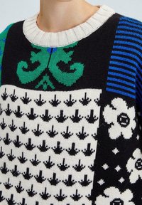 Pull en tricot présentant plusieurs motifs : fleurs vertes, flèches noires et blanches, rayures bleues. Encolure côtelée crème ; texture douce.