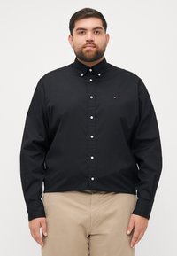 Black button-up shirt with long sleeves, featuring a pointed collar and white buttons, made from smooth fabric. Small logo on the chest.
