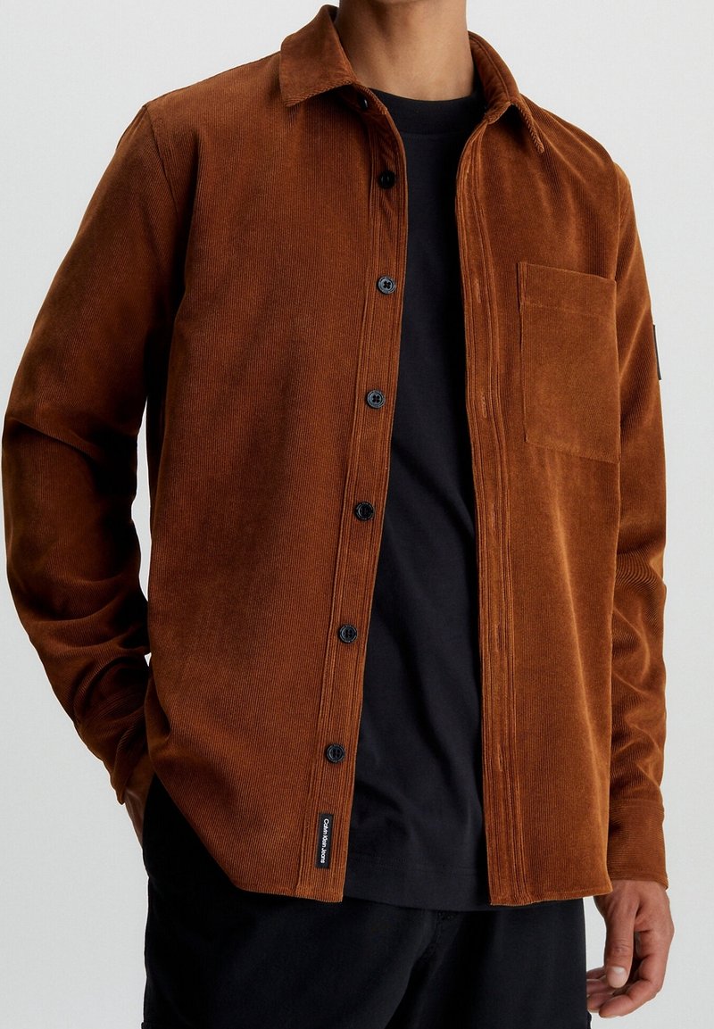 Camicia button-up in corda marrone con colletto, taschino frontale e bottoni neri, indossata sopra una t-shirt nera. Tessuto testurizzato e caldo.