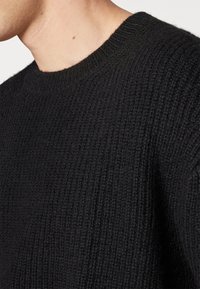 Svart stickad tröja med ribbad textur och rund halsringning. Tyget ser mjukt och mysigt ut, med en närbild av sömmarna.