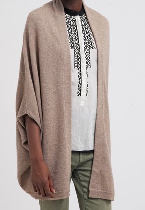 Personne portant un châle beige ouvert à l'avant sur une chemise blanche avec une broderie géométrique noire et un pantalon vert olive.