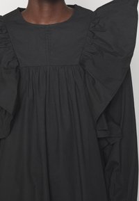 Mørkt stofkjole med brede, flæseformede skuldre og sammensyede plisseringer under en firkantet bærestykke på en person med mørk hud.