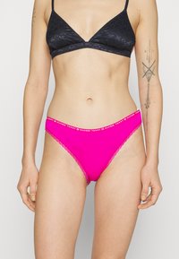 Rozā bikini apakšbikses ar mežģīņu apdari, zīmola jostu un gludu tekstūru. Apvienotas ar melnu rakstainu braleti.