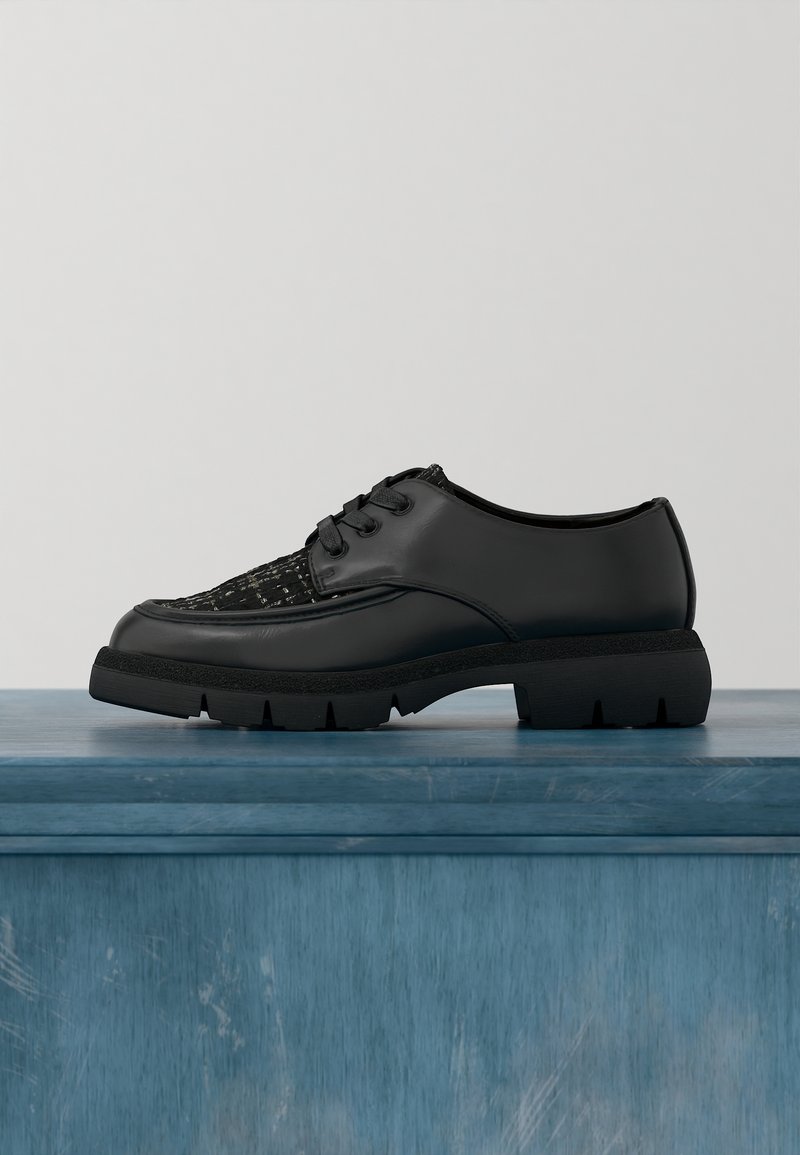 Chaussure noire en cuir à lacets avec panneau avant texturé et semelle épaisse à crampons, présentée sur une surface en bois bleue sur un fond uni.