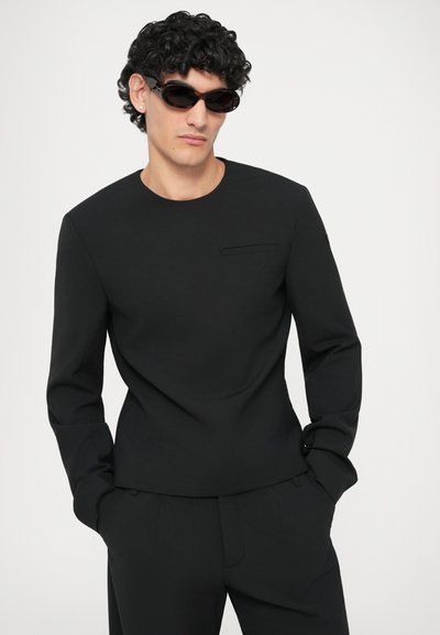 Tricou negru cu mânecă lungă și guler rotund, având o mică buzunar pe piept. Purcat cu ochelari de soare, arătând un material neted și structurat.
