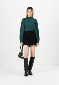Zelená halenka s dlouhým rukávem a napufnutými rameny, spárovaná s černou mini sukni s bočními rozparky, černými vysokými botami po kolena a malou kabelkou.