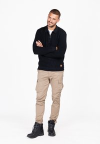 Schwarzer Strickpullover mit Reißverschlusskragen, beigen Cargo-Hosen und schwarzen Schnürstiefeln. Der Pullover hat ein kleines braunes Markenetikett am Saum.