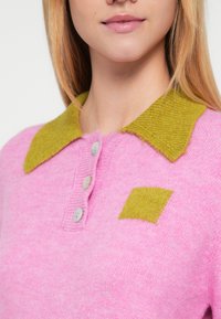 Maglione rosa lavorato a maglia con colletto verde testurizzato e dettaglio quadrato verde. Presenta una chiusura con bottoni al collo. Materiale morbido.