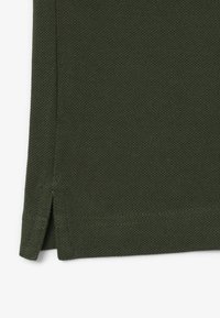 Gros plan d’un tissu texturé vert foncé avec une fente latérale et des coutures visibles sur l’ourlet et la couture.