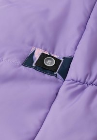 Tela acolchada morada con una textura suave y una etiqueta de tela con un patrón floral que presenta un cierre de Velcro negro y un detalle de botón transparente.