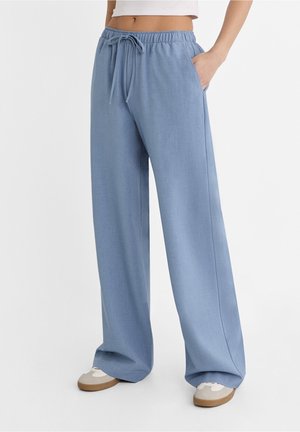 Blaue Hosen mit weitem Bein und Kordelzug in der Taille, getragen mit weißen Sneakers und einem weißen, kurz geschnittenen Oberteil, in Pose mit der Hand in der Tasche.