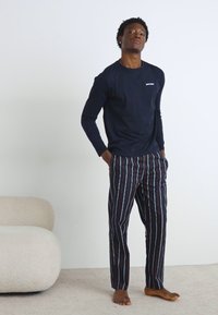 Mørkeblå skjorte med lange ærmer og hvidt logo, parret med flerfarvede stribede bukser i mørke nuancer, der har lige ben og uden synlige sko.