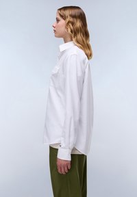 Camicia bianca a maniche lunghe con colletto alla francese, dotata di taschino sul petto e vestibilità ampia, abbinata a pantaloni verdi. Tessuto morbido.