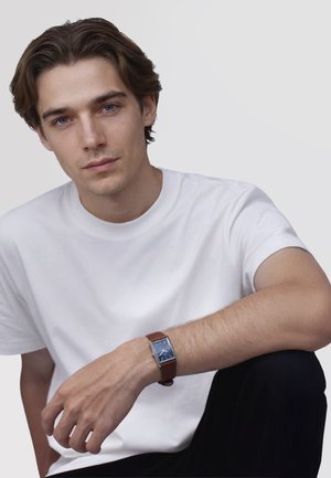 Fiatal férfi fehér pólóban, barna bőrszíjas, téglalap alakú kék számlapos karórát visel, egyszínű világos háttér előtt ül.