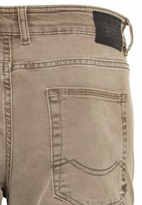 Los pantalones de mezclilla color canela cuentan con un bolsillo trasero con detalles de costura y una etiqueta de marca de cuero negro en la cintura. Textura suave, diseño clásico.