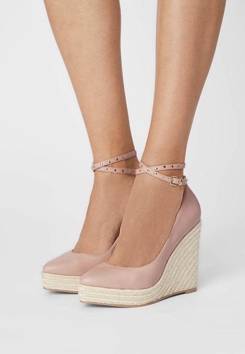 Jambes portant des espadrilles compensées roses avec des brides à la cheville et une boucle dorée, semelle plateforme tissée naturelle, debout sur une surface blanche.