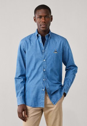 Man wearing light blue denim button-up shirt over dark t-shirt and beige pants, standing against plain light background.
