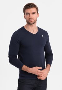 Laivan tummansininen pitkähihaisella V-pääntiellä oleva paita, joka on valmistettu pehmeästä kankaasta ja siinä on kapea muotoilu. Paidassa on pieni logo rinnassa ja suora helma.