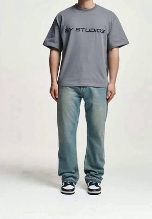 Personne portant un T-shirt gris oversize avec le texte "2Y STUDIOS", un jean bleu ample et des baskets noires et blanches, debout sur un fond uni.