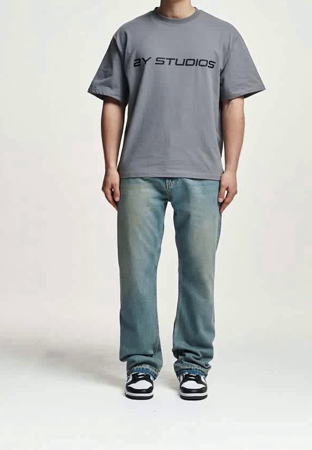 Personne portant un T-shirt gris oversize avec le texte "2Y STUDIOS", un jean bleu ample et des baskets noires et blanches, debout sur un fond uni.