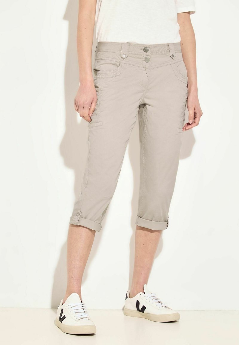 Beige vaatteet, joissa on käännetyt lahkeet, etutaskut ja kaksoispainonappi kiinnitys. Sileä rakenne, rento muotoilu.