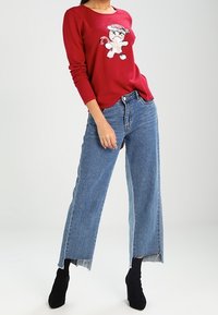 Personne portant un haut rouge à manches longues avec un chat cartoon, un jean taille haute bleu aux ourlets effilochés, associé à des bottines noires à talons.