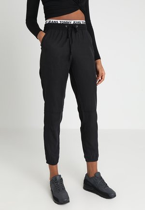 Person trägt schwarze Jogginghosen mit elastischen Bündchen, weißen Bund mit Aufschrift "Tommy Jeans", schwarzes Crop-Top und dunkelgraue Nike-Sneaker.