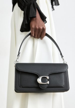 Mørk hudfarvet hånd, der holder en sort læder håndtaske med en sølv "C" spænde mod hvide klæder og en sort rynketærme.