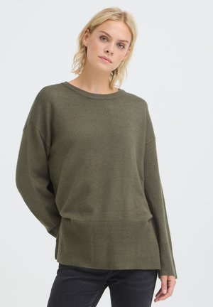 Femme aux cheveux blonds courts portant un pull vert olive oversized et un pantalon noir sur un fond clair uni.