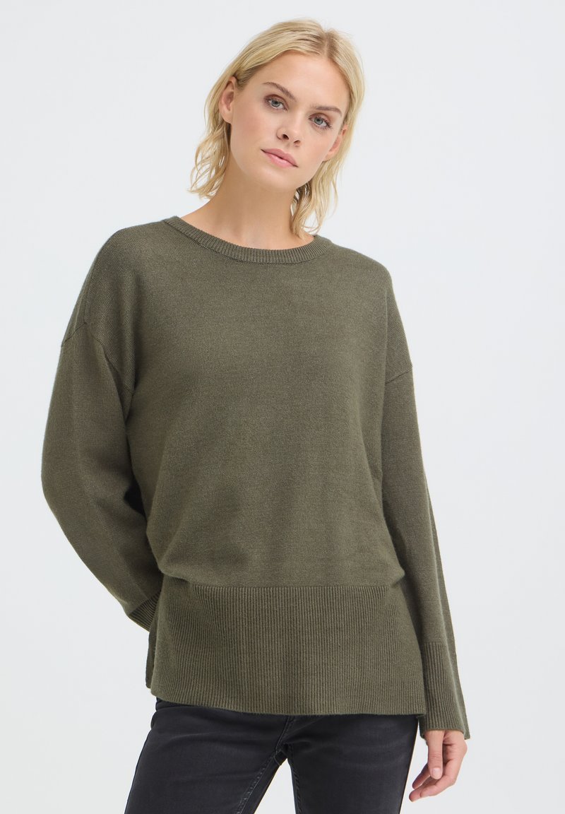 Femme aux cheveux blonds courts portant un pull vert olive oversized et un pantalon noir sur un fond clair uni.